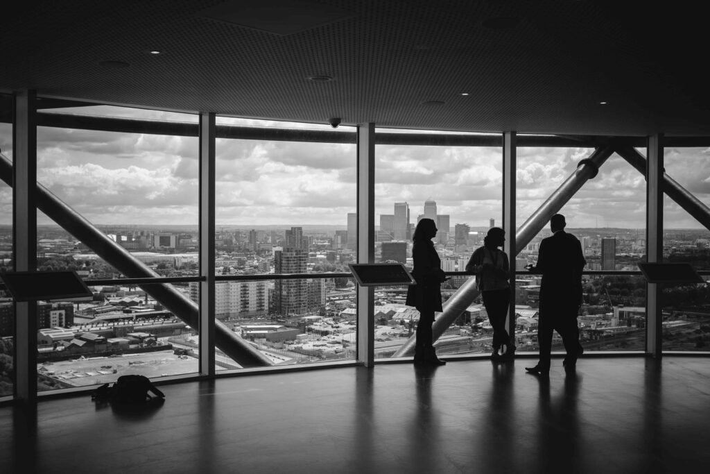 Black and white photo of three people silhouetted against a wall of windows above a city