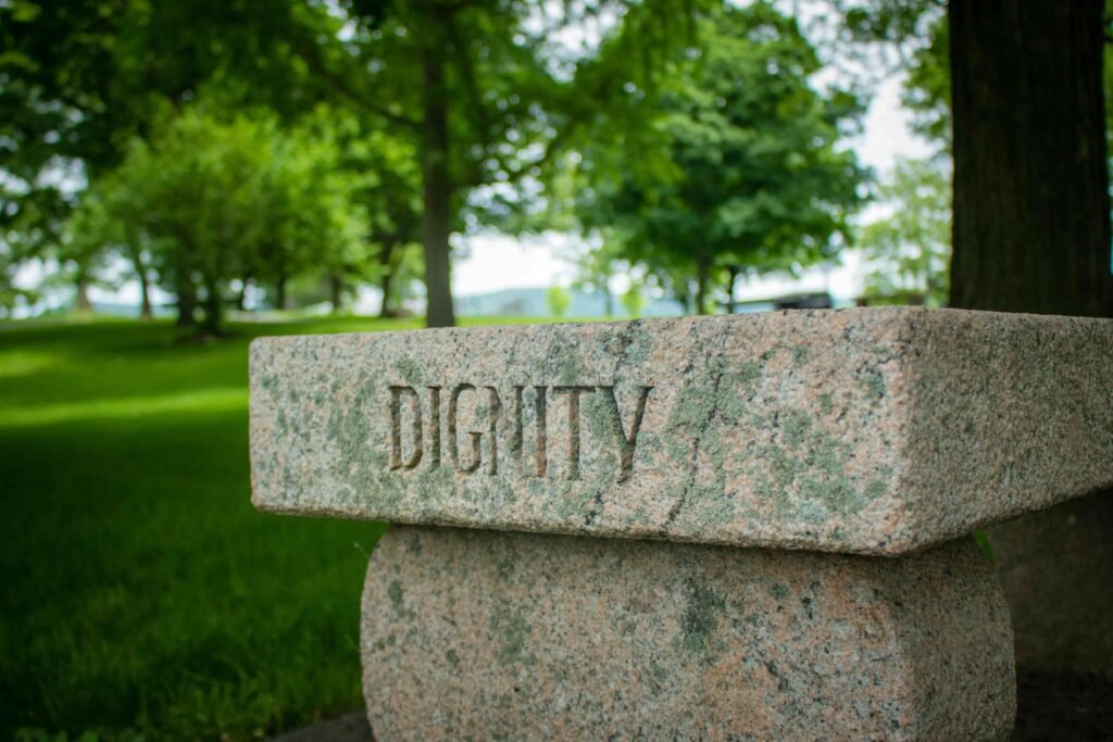 A granite stone carved with the word Dignity, on a green lawn beneath trees