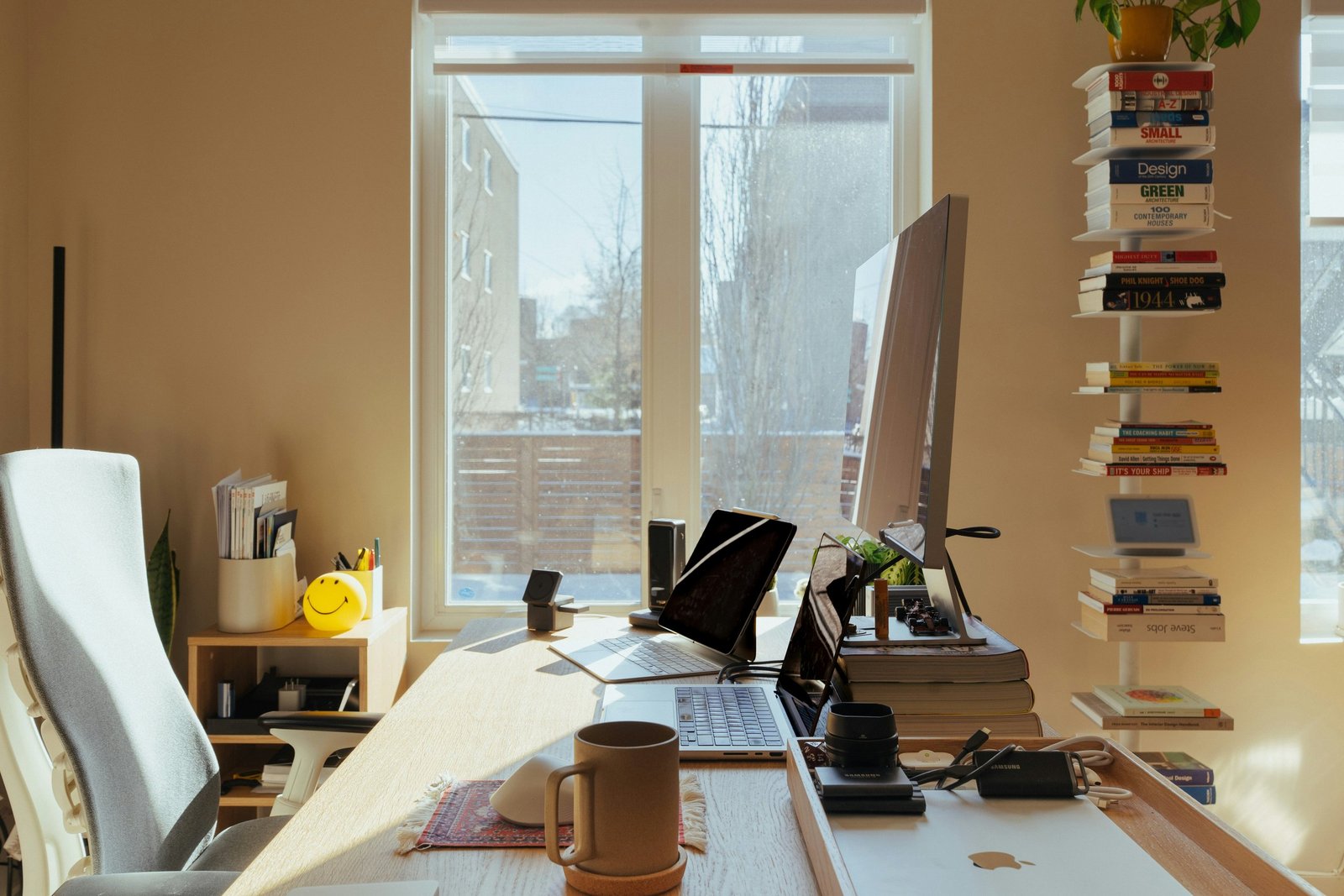 Sunlit home office with a timber desk, monitor, laptops and a coffee mug, and a tall stack of books by the window